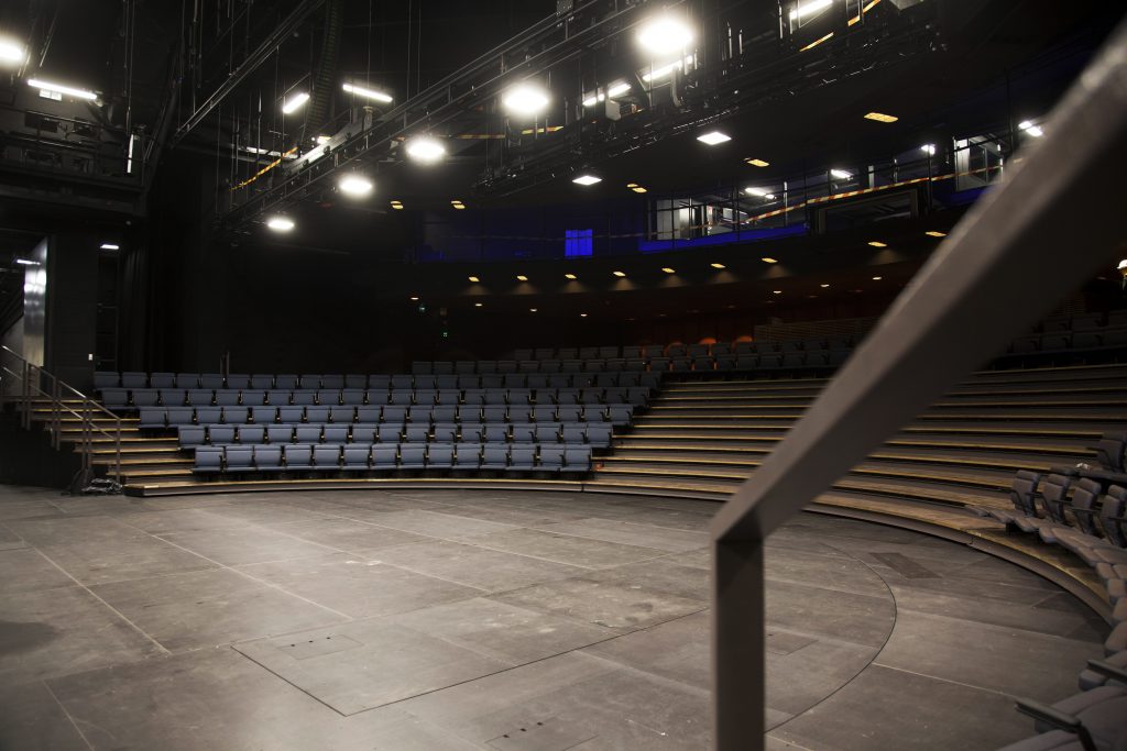 Näkymä tyhjän, modernin teatterin näyttämöltä, jossa on porrastetut istuimet, kaarevat sinisten tuolien rivit ja kirkkaat näyttämövalot, jotka valaisevat tilaa.