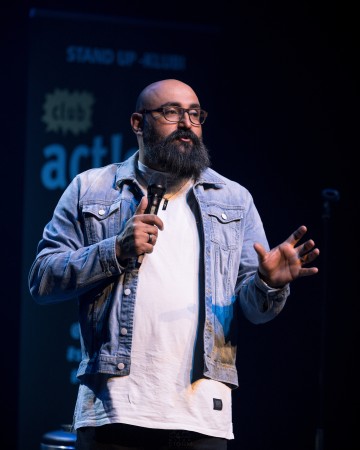 Silmälasipäinen ja farkkutakkinen parrakas mies pitelee mikrofonia esiintyessään stand up -komiikkaa lavalla, ja hänen takanaan on sumea kyltti ja tumma tausta.