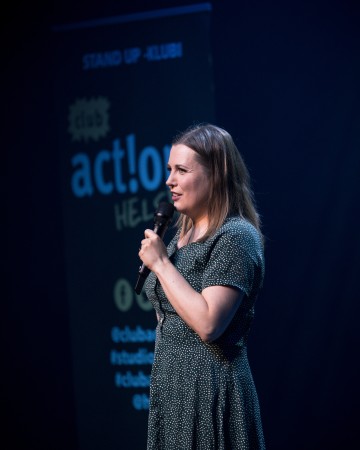 Mikrofoni kädessään pitävä nainen esittää stand up -komiikkaa lavalla, ja taustalla näkyy sumea tapahtumabanneri.