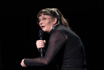 Silmälasipäinen henkilö, jolla on pitkät ruskeat hiukset poninhännässä, pitää lavalla mikrofonia, hänellä on yllään läpikuultava musta toppi ja suuret korvakorut, ja hän puhuu vilkkaasti tummaa taustaa vasten.