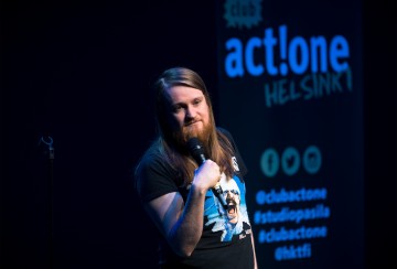 Pitkätukkainen parrakas koomikko esiintyy lavalla mikrofoni kädessä, taustalla sininen act!one HELSINKI -kyltti ja sosiaalisen median kahvat.
