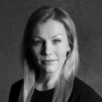 Mustavalkoinen muotokuva naisesta, jolla on suorat, vaaleat hiukset, yllään tumma toppi, katselee suoraan kameraan neutraali ilme tavallista taustaa vasten.