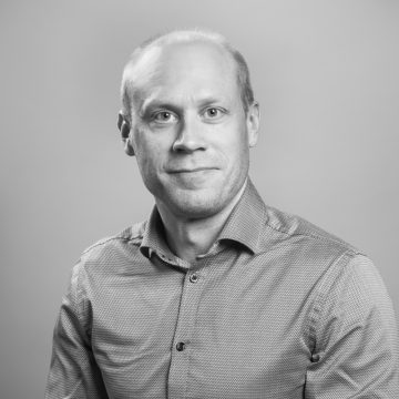 Lyhyet, vaaleat hiukset ja hento hymy on miehellä, jolla on kuvioitu napitettava paita. Hän katsoo kameraan vaaleaa taustaa vasten mustavalkoisessa muotokuvassa.