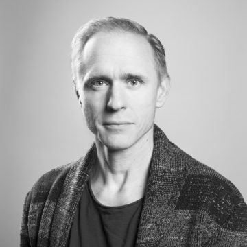 Mustavalkoinen muotokuva keski-ikäisestä miehestä, jolla on lyhyet vaaleat hiukset, päällään kuviollinen neuletakki ja tumma t-paita, ja joka katsoo suoraan kameraan neutraali ilme taustaa vasten.
