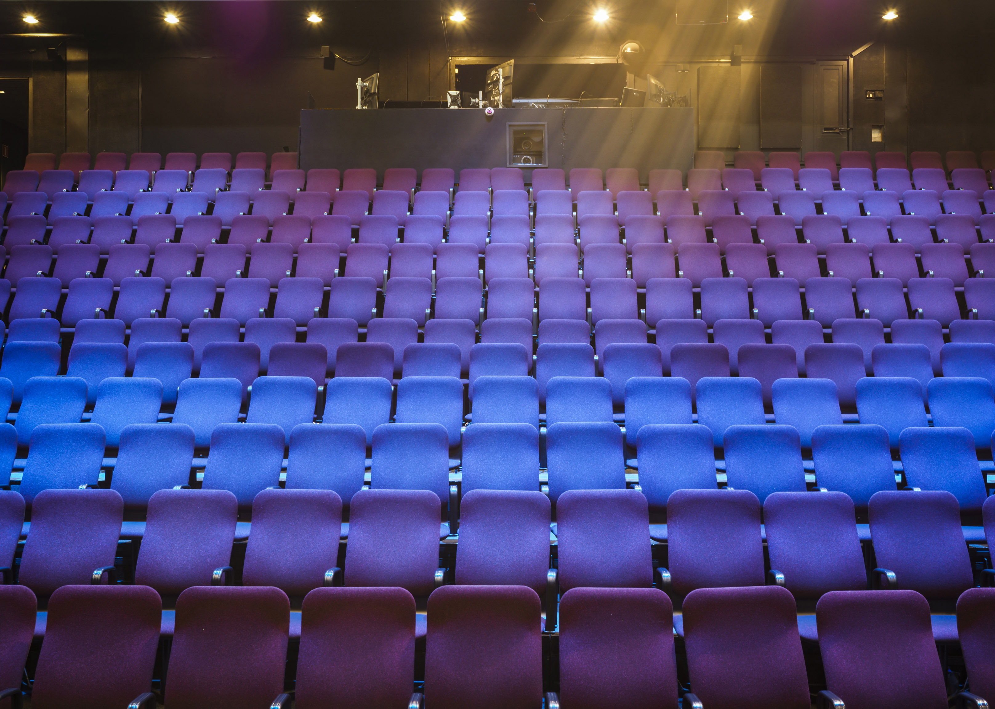 Tyhjien violettien ja sinisten teatteripenkkien rivistöistä on edessä takana oleva ohjauskoppi, jota valaisee muutama yläpuolella oleva valonheitin muuten hämärässä salissa.