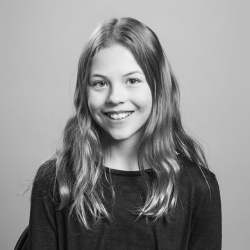 Nuori tyttö, jolla on pitkät, aaltoilevat hiukset ja musta toppi, hymyilee kameralle. Kuva on mustavalkoinen, ja siinä on tavallinen tausta.