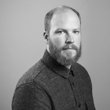 Mustavalkoinen muotokuva parrakas mies, jolla on lyhyet hiukset, päällään napitettu, kuvioitu tumma paita, katselee suoraan kameraan tavallista taustaa vasten.