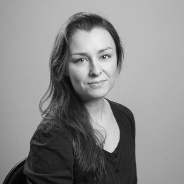Nainen, jolla on pitkät tummat hiukset ja tumma, napitettu paita, istuu tuolilla ja hymyilee hieman kameralle. Tausta on tavallinen ja vaalea. Kuva on mustavalkoinen.