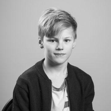 Mustavalkoinen muotokuva vaaleahiuksisesta nuoresta pojasta, jolla on vaalean paidan päälle puettu tumma villatakki, joka istuu tuolilla ja katsoo neutraalilla ilmeellä kameraan tavallista taustaa vasten.