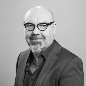 Keski-ikäinen mies, jolla on kalju pää, parta ja silmälasit, yllään puvun takki ja tumma paita, hymyilee hieman vaaleanharmaata taustaa vasten.