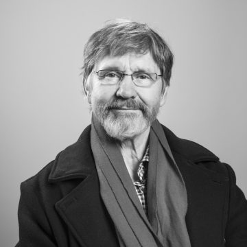 Mustavalkoinen muotokuva vanhemmasta miehestä, jolla on silmälasit, parta ja viikset. Hänellä on takki ja huivi, hän hymyilee lempeästi ja katsoo suoraan kameraan tavallista taustaa vasten.