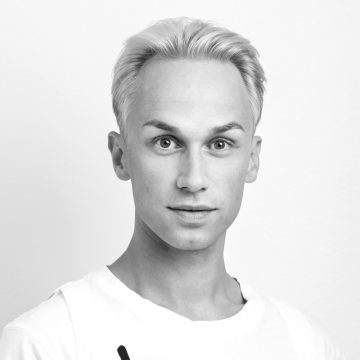 Mustavalkoinen muotokuva henkilöstä, jolla on vaalea iho ja lyhyet vaaleat vaaleat hiukset, yllään tavallinen valkoinen toppi, katselee suoraan kameraan neutraali ilme tavallista taustaa vasten.