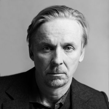 Keski-ikäinen mies, jolla on vaaleat hiukset ja lyhyt parta, katsoo suoraan kameraan. Hänellä on tumma puvuntakki ja vakava ilme, ja hän on asettunut tavallista vaaleaa taustaa vasten. Kuva on mustavalkoinen.