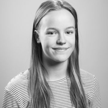 Mustavalkoinen muotokuva nuoresta henkilöstä, jolla on suorat, pitkät hiukset, yllään raidallinen paita, hymyilee pehmeästi ja katsoo kameraan tavallista taustaa vasten.