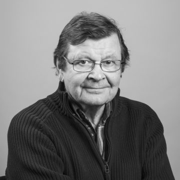 Vanhempi mies, jolla on silmälasit ja lyhyet tummat hiukset, istuu kameraan päin. Hänellä on vetoketjullinen villapaita ruudullisen paidan päällä. Tausta on tavallinen ja vaalea, ja kuva on mustavalkoinen.