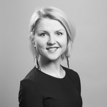 Mustavalkoinen muotokuva hymyilevästä naisesta, jolla on lyhyet vaaleat hiukset, pitkät korvakorut ja tumma toppi, ja joka katsoo suoraan kameraan tavallista taustaa vasten.