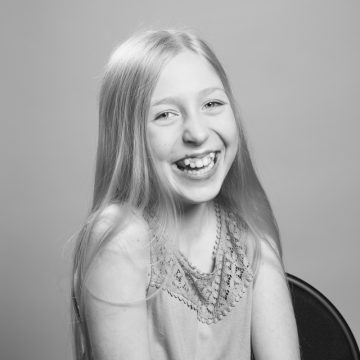 Nuori tyttö, jolla on pitkät, suorat hiukset, hymyilee leveästi ja näyttää hampaansa. Hänellä on yllään hihaton toppi, jossa on pitsikuvio, ja hän istuu tuolilla yksinkertaista taustaa vasten. Kuva on mustavalkoinen.