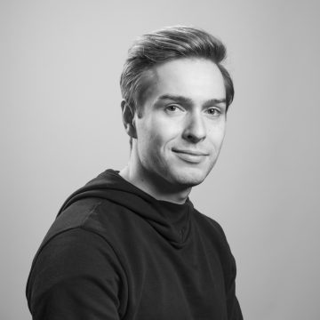Mustavalkoinen muotokuva vaaleahiuksisesta nuoresta miehestä, jolla on tumma huppari päällään, katselee kameraan ja hymyilee hieman, taustana tavallinen tausta.