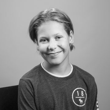 Tässä mustavalkoisessa muotokuvassa vaaleahiuksinen nuori henkilö, jolla on yllään tumma paita, jossa on ympyränmuotoinen tunnus, istuu ja hymyilee kameralle tavallista taustaa vasten.