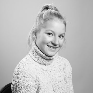 Mustavalkoinen muotokuva nuoresta henkilöstä, jolla on vaaleat hiukset poninhännässä, päällään kuvioitu poolopusero, hymyilee ja katsoo hieman sivulle tavallista taustaa vasten.