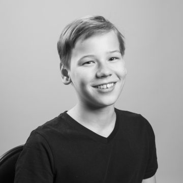 Hymyilevä nuori poika, jolla on lyhyet hiukset ja musta V-kaula-aukkoinen paita, istuu tuolilla vaaleaa taustaa vasten. Kuva on mustavalkoinen.