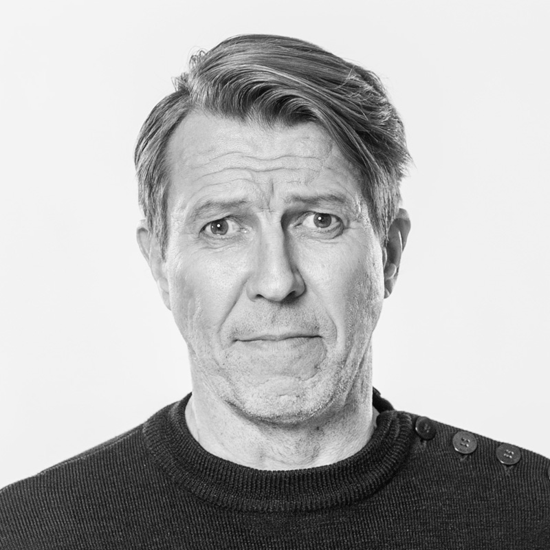 Mustavalkoinen muotokuva keski-ikäisestä, vaaleahiuksisesta miehestä, jolla on tumma villapaita, jonka olkapäässä on napit, ja joka katsoo suoraan kameraan hieman huolestunut tai epävarma ilme.