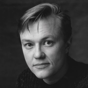 Mustavalkoinen muotokuva henkilöstä, jolla on lyhyet, vaaleat hiukset, yllään tumma pusero, katselee suoraan kameraan neutraali ilme tummalla taustalla.