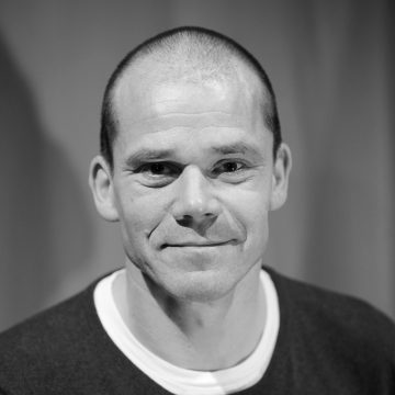 Mustavalkoinen muotokuva miehestä, jolla on ajeltu pää, yllään tumma villapaita valkoisen paidan päällä, hymyilee hellästi ja katsoo suoraan kameraan neutraalilla taustalla.