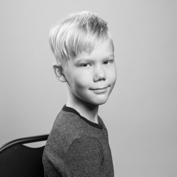 Mustavalkoinen muotokuva nuoresta pojasta, jolla on lyhyet vaaleat hiukset ja raidallinen paita, joka istuu sivuttain tuolilla ja katsoo kameraan hymyillen. Tasainen tausta.