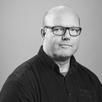 Mustavalkoinen muotokuva keski-ikäisestä henkilöstä, jolla on lyhyet hiukset ja silmälasit, yllään napillinen paita, katselee suoraan kameraan neutraali ilme taustaa vasten.