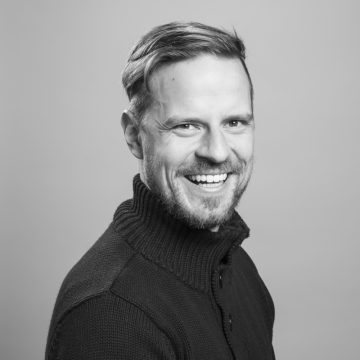Lyhyet, vaaleat hiukset ja partainen mies hymyilee leveästi ja pukeutuu tummaan, korkeakauluksiseen puseroon. Kuva on mustavalkoinen ja taustaltaan tavallinen.