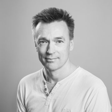 Mies, jolla on lyhyet, piikikkäät hiukset ja vaalea, napitettu paita, poseeraa ja hymyilee pehmeästi kameralle vaaleaa taustaa vasten. Kuva on mustavalkoinen.