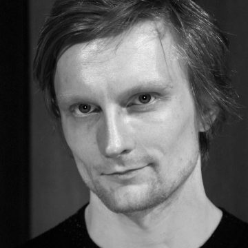 Mustavalkoinen muotokuva henkilöstä, jolla on lyhyet, kiharat hiukset, vaalea iho ja hento hymy, päällään tumma toppi ja joka katsoo suoraan kameraan epäselvää taustaa vasten.