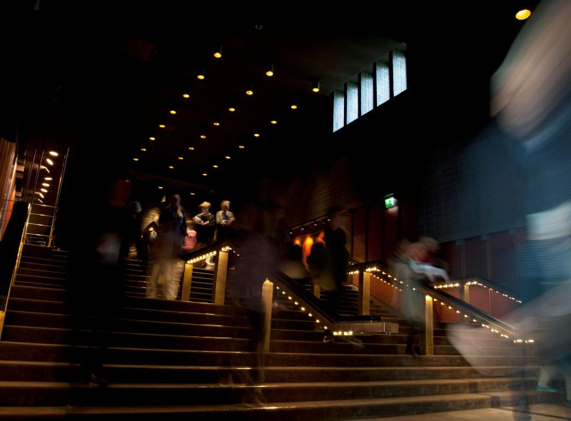 Hämärästi valaistu teatterin tai elokuvateatterin portaikko, jossa hämäriä hahmoja liikkuu ylös- ja alaspäin ja jota valaisevat pienet valot käsijohteissa ja portaissa.
