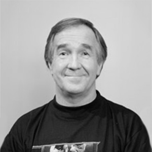 Keski-ikäinen mies, jolla on lyhyet vaaleat hiukset, hymyilee hieman ja jolla on tumma t-paita yllään. Tausta on tavallinen ja vaalea. Kuva on mustavalkoinen.