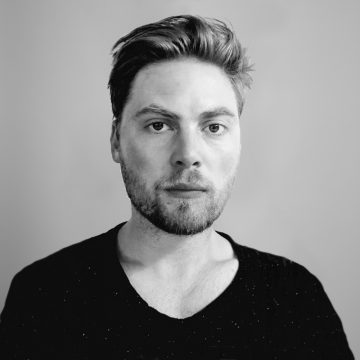 Mustavalkoinen muotokuva miehestä, jolla on lyhyet, muotoillut hiukset ja leikattu parta, tumma paita päällään ja joka katsoo suoraan kameraan neutraali ilme kasvoillaan tavallista taustaa vasten.
