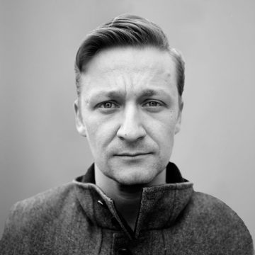Mies, jolla on lyhyet vaaleat hiukset ja vakava ilme, katsoo suoraan kameraan. Hänellä on napitettu takki ja hän on poseerannut vaaleaa taustaa vasten.