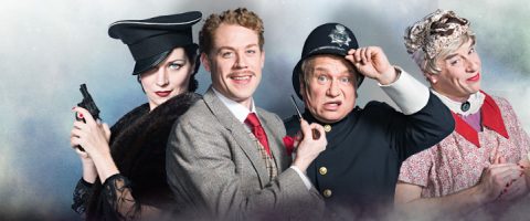 Neljä teatteripukuista henkilöä: mustahattuinen nainen, jolla on ase kädessään, pukuinen mies, joka hymyilee, poliisin univormuun pukeutunut mies, joka näyttää yllättyneeltä, ja iäkkääksi naiseksi pukeutunut mies, jolla on harmaat kiharat ja vaaleanpunainen mekko.