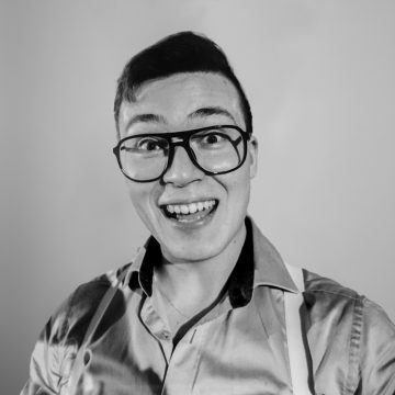 Nuori henkilö, jolla on suuret silmälasit ja napillinen paita, hymyilee laajasti kameralle tavallista taustaa vasten. Kuva on mustavalkoinen.