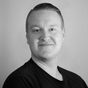 Mies, jolla on lyhyet, liukuhiuksiset hiukset ja hento hymy, poseeraa vaaleaa taustaa vasten. Hänellä on yllään tumma t-paita ja hän katsoo hieman oikealle.