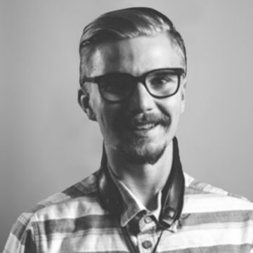 Mustavalkoinen muotokuva lyhythiuksisesta, silmälasipäisestä ja viiksekkäästä nuoresta miehestä, jolla on raidallinen paita ja kuulokkeet kaulassaan ja joka hymyilee kameralle tavallista taustaa vasten.
