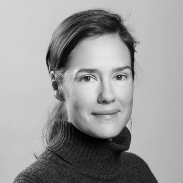 Mustavalkoinen muotokuva naisesta, jolla on vaalea iho ja tummat hiukset taaksepäin vedettynä, päällään uurrettu villapaita, katselee kameraan ja hymyilee pehmeästi tavallista taustaa vasten.