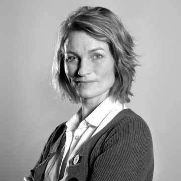 Vaaleat, polkkatukkainen nainen, jolla on kauluspaita ja villatakki, poseeraa tavallista taustaa vasten ja katsoo luottavaisesti kameraan mustavalkoisena.