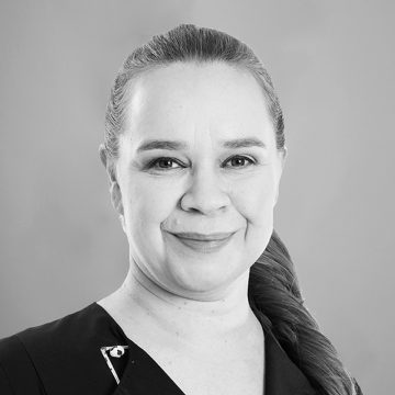 Vaaleaihoinen nainen, jolla on pitkät, poninhännäksi taakse sidotut hiukset, hymyilee lempeästi kameralle. Hänellä on yllään tumma toppi, jossa on pieni koristepinssi. Tausta on tavallinen ja vaalea.