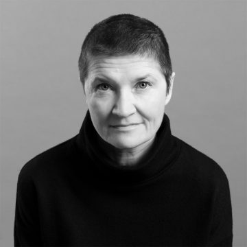 Keski-ikäinen lyhythiuksinen henkilö, jolla on tumma poolopusero, katsoo suoraan kameraan vaaleanharmaalla taustalla. Ilme on neutraali ja kuva on mustavalkoinen.