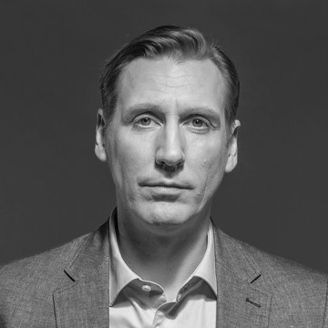 Mustavalkoinen muotokuva miehestä, jolla on puvuntakki ja avokauluspaita ja joka katsoo neutraalilla ilmeellä suoraan kameraan tavallista taustaa vasten.