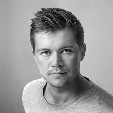 Nuorella miehellä, jolla on lyhyet, hieman sotkuiset hiukset ja vaaleat karvat, on yllään vaalea t-paita, ja hän katsoo neutraalilla ilmeellä suoraan kameraan tavallista taustaa vasten.