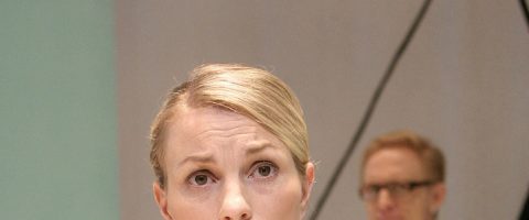Etualalla seisoo nainen, jolla on taaksepäin vedetyt vaaleat hiukset ja joka näyttää huolestuneelta, kun taas taustalla on sumea silmälasipäinen mies.