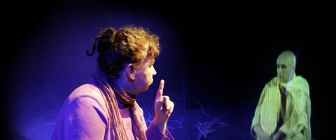 Purppuranpunaiseen puseroon ja huiviin pukeutunut nainen nostaa sormeaan ja katsoo istuvaa hahmoa, joka näyttää heijastuvan hologrammina tummaa taustaa vasten.