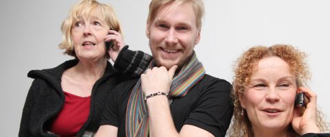 Kolme ihmistä seisoo vierekkäin hymyillen. Kaksi naista, toisella lyhyet vaaleat hiukset ja toisella vaaleat kiharat hiukset, puhuvat kännykkään. Heidän välissään seisoo vaaleahiuksinen, huiviin pukeutunut mies, joka hymyilee kameralle.